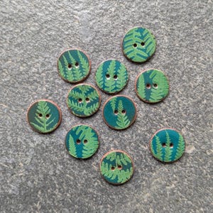 May include: Ten round buttons with a fern leaf pattern. The buttons feature a teal base with green fern leaves and a copper-coloured edge. Each button has two holes for sewing.