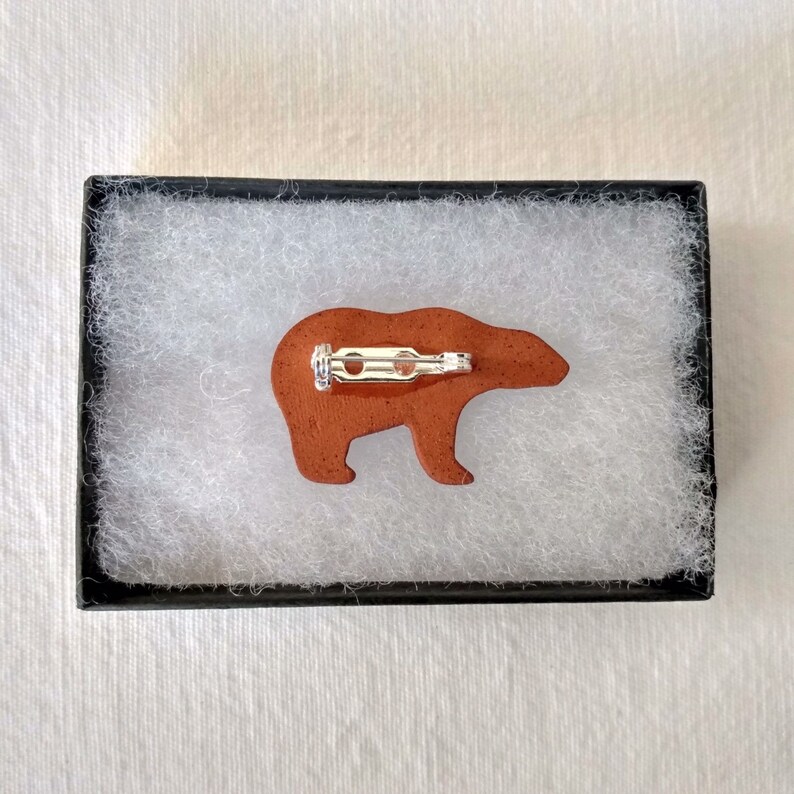 May include: A brown ceramic bear pin with a silver pin back. The bear is facing to the right.