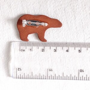 May include: A brown leather bear pin with a metal clasp. The pin is approximately 3 cm long and 2 cm wide.
