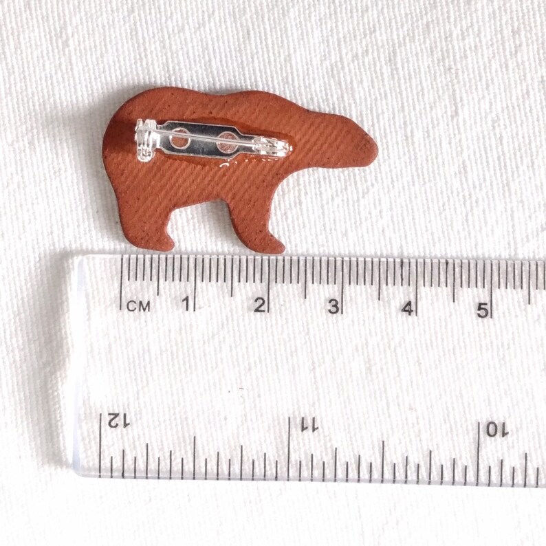 May include: A brown leather bear pin with a metal clasp. The pin is approximately 3 cm long and 2 cm wide.