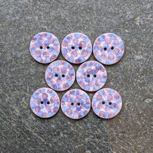 May include: A set of eight round buttons featuring a speckled pattern of light blue and reddish-purple dots. Each button is circular with two small holes in the centre for attachment. The surface of the buttons has a textured, matte appearance.