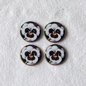 May include: Four round buttons with a floral design. Each button features a white pansy flower with purple and yellow accents, set against a green background. The buttons are arranged on a white fabric surface.