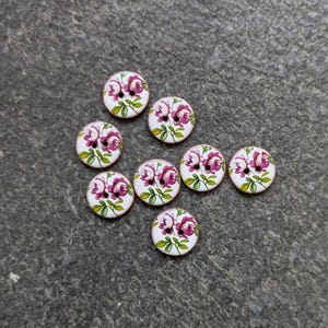 Op de afbeelding: Een verzameling van negen ronde houten knopen met een bloemenontwerp. Elke knoop heeft een witte achtergrond met een roze roos en groene bladeren. De knopen zijn verspreid over een grijze ondergrond.