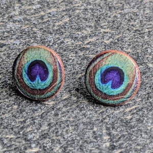 Puede incluir: Dos pendientes de botón redondos con diseño de pluma de pavo real. Cada pendiente presenta un óvalo central morado rodeado de círculos concéntricos turquesa, marrón y rojo. Sobre una superficie gris texturizada.
