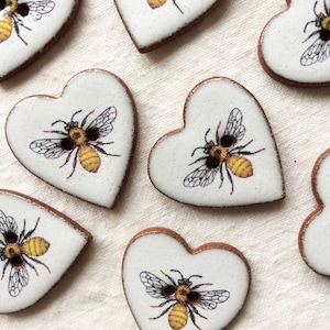 May include: A set of ten heart-shaped ceramic buttons with a white background and a brown border. Each button features a detailed illustration of a bee with yellow and black stripes.