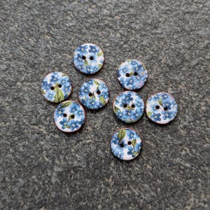 May include: Eight round buttons with a floral pattern. Each button has a white base with blue flowers and green leaves. The buttons have two holes for sewing and a brown edge. Ideal for sewing projects.