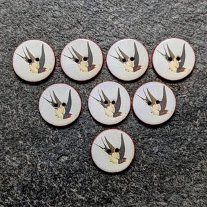 May include: Seven circular, off-white buttons, each featuring a two-hole design and a printed illustration of a flying swallow with black wings and a cream-coloured chest. The buttons are arranged in a triangular formation, displaying a consistent bird motif.
