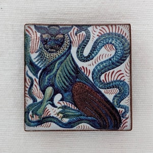 Puede incluir: Un azulejo de cerámica con una criatura mítica azul y verde con una cola roja y marrón. La criatura está rodeada por una serpiente azul y remolinos rojos y blancos.