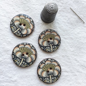May include: Four round buttons featuring a repeating floral pattern in shades of yellow, peach, and black, each with two central holes. A metallic silver thimble with a textured surface and a single thin sewing needle are positioned nearby.