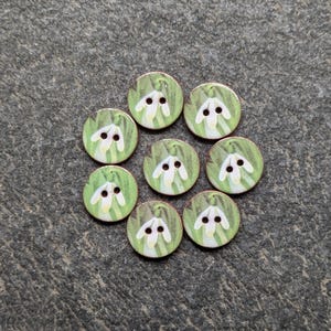 Op de afbeelding: Acht ronde houten knopen met een geschilderd sneeuwklokjesontwerp. Elke knoop heeft een witte bloem met groene bladeren op een groene achtergrond. De knopen zijn in een cirkel gerangschikt op een grijze ondergrond.