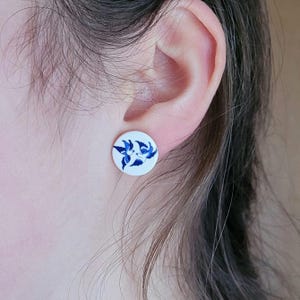 Könnte beinhalten: Weiße, runde Ohrstecker mit blauem Blumenmuster. Die Ohrringe sind vor dem Hintergrund eines Ohrs und der Haut einer Person platziert, wodurch die Details des Schmucks hervorgehoben werden. Das Blau-Weiß-Farbschema verleiht den Ohrringen ein klassisches Aussehen.