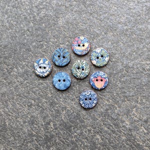 May include: A collection of eight decorative buttons with floral and bird patterns. The buttons are primarily blue, with accents of pink, orange, and green. Each button has two holes for sewing.