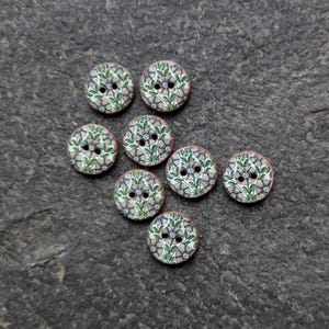 May include: Eight round wooden buttons with a floral design. Each button has a pattern of green leaves and grey flowers on a white background. The buttons have two holes for sewing and a reddish-brown edge.