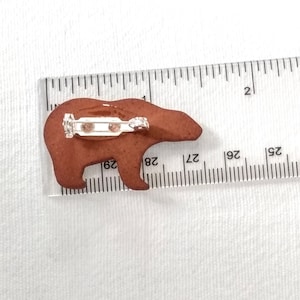 May include: A brown ceramic bear pin with a silver pin back. The bear is about 1 inch tall and 1.5 inches wide.