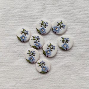 May include: Eight white, circular buttons, each adorned with a hand-painted design of blue forget-me-not flowers and green leaves. The buttons are arranged on a white, textured fabric surface.