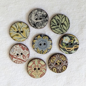 May include: Assortment of eight round, fabric-covered buttons with various floral and nature-inspired designs. The buttons are in shades of brown, green, blue, and white.
