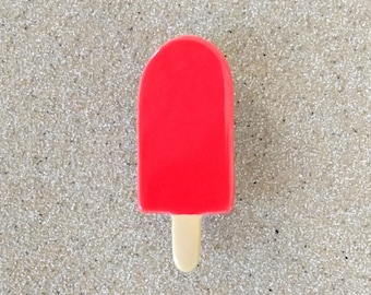 May include: A bright red popsicle-shaped object with a smooth, glossy finish and a small, light beige wooden stick protruding from the bottom. The item features a rounded top edge and a flat, rectangular body, presenting a simple, solid color design.