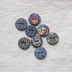 Op de afbeelding: Acht ronde houten knopen met bloemen- en vogelpatronen. De knopen hebben een donkerblauwe achtergrond met patronen in blauw, groen en crème. Elke knoop heeft twee of vier gaten om op te naaien. Diameter ongeveer 2,5 cm.