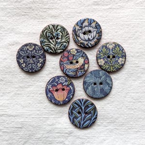 May include: Eight round wooden buttons with diverse floral and bird patterns. The buttons have a dark blue base with designs in green, pink, and white. Each button is designed with two holes for sewing, perfect for craft projects.