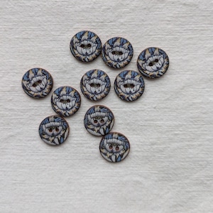 Peut inclure: Un ensemble de dix boutons ronds en bois, chacun présentant un motif floral détaillé dans des tons de bleu, blanc et or. Chaque bouton possède deux trous centraux pour la fixation et un bord couleur bois naturel, disposés en vrac.