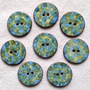 May include: A set of eight circular buttons featuring a speckled pattern in shades of blue, green, and yellow. Each button has a smooth, glossy finish and two central holes for attachment, arranged in a symmetrical pattern on a textured surface.