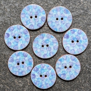 May include: Seven round buttons featuring a speckled pattern of light blue, periwinkle, and white dots. Each button has two central holes and a smooth, circular edge. The surface of the buttons displays a mottled, multi-coloured finish.