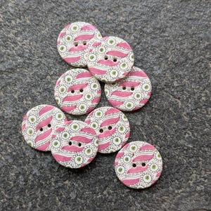May include: A collection of eight round, wooden buttons with a pink and green floral pattern. The buttons are approximately 1 inch in diameter.