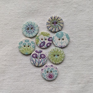 May include: A collection of eight round wooden buttons with floral designs in various colors. The buttons feature patterns in shades of blue, green, purple, and white. Each button has two holes for sewing.