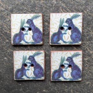 May include: Four square wooden buttons featuring a design of two blue rabbits on a cream background with a gold leaf pattern. Each button has two holes for sewing and a reddish-brown border.
