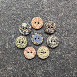May include: Eight round wooden buttons with diverse floral and patterned designs. The buttons display colours such as blue, orange, yellow, and green. Each button has two holes for sewing and is arranged in a circular pattern.