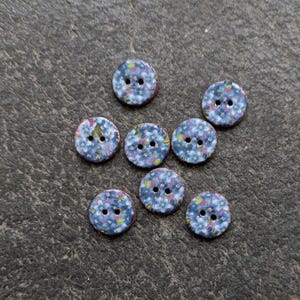 May include: Seven round buttons with a floral design. The buttons feature a blue base with small white and colourful flower details. Each button has two sewing holes and a brown edge. Ideal for sewing projects.