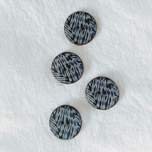 May include: Four circular buttons with a dark blue and white patterned design. Each button has two holes for sewing. The buttons are arranged on a white fabric surface.