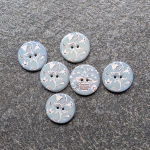 May include: Six round, light blue buttons with a floral design. Each button features a white floral pattern and two holes for sewing. The buttons are arranged on a gray textured surface.