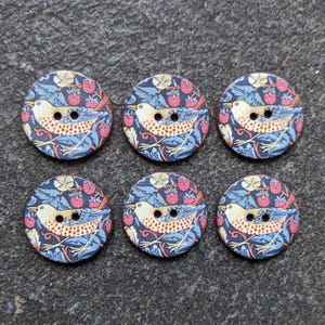 Puede incluir: Seis botones redondos de madera con un diseño de pájaro y bayas. Los botones presentan un pájaro color crema con detalles marrones, rodeado de bayas rojas y hojas azules sobre un fondo azul marino. Cada botón tiene dos agujeros para coser.