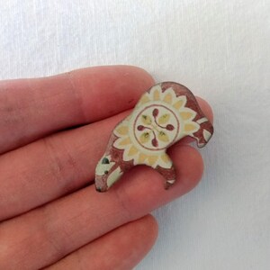 May include: A small, ceramic bear figurine with a red, yellow, and white floral pattern. The bear is standing on its hind legs and has a round, floral design on its chest.