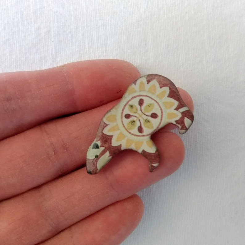 May include: A small, ceramic bear figurine with a red, yellow, and white floral pattern. The bear is standing on its hind legs and has a round, floral design on its chest.