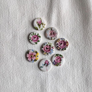 May include: A collection of seven round buttons featuring various floral patterns. Each button displays distinct botanical illustrations including pink roses, rosebuds, and small blue flowers on white backgrounds, with two holes in the centre of each piece.