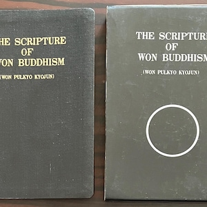 Puede incluir: Dos libros negros con letras doradas. El texto en las portadas dice "THE SCRIPTURE OF WON BUDDHISM" y "(WON PULKYO KYOJUN)". Un libro está abierto, el otro está en una caja con un círculo blanco.