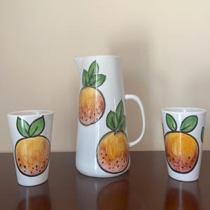 Puede incluir: Una jarra de cerámica blanca y dos vasos a juego. Cada pieza está decorada con un diseño de fruta de naranja pintado a mano con hojas verdes. La jarra tiene un asa y es más alta que los vasos. El juego se exhibe sobre una superficie marrón oscuro.