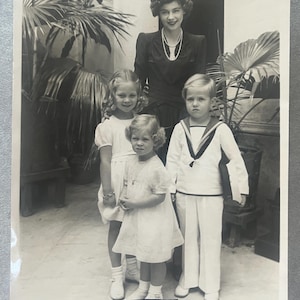 May include: A vintage black and white photograph showing a woman and three children posing outside. The woman wears a dark dress and a pearl necklace. The children are in white outfits. A signature is at the bottom.