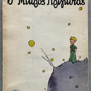 May include: Vintage book cover featuring the title "O Mikros Prigkips" in Greek script, with the author's name, Antoine de Saint-Exupéry. The illustration shows a small figure in green on a planet with stars and planets.