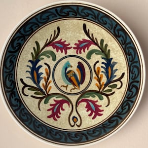 Puede incluir: Un plato de cerámica vintage con fondo crema y un diseño de pájaro estilizado en azul, verde, naranja y rojo. El plato tiene un borde azul oscuro y negro con un patrón repetitivo.