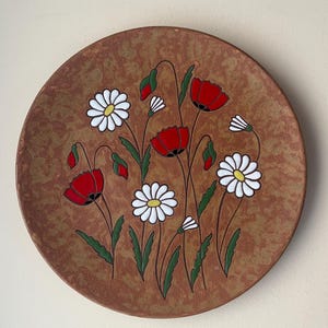 Può includere: Un piatto in terracotta con un disegno dipinto a mano di papaveri rossi e margherite bianche su uno sfondo marrone.
