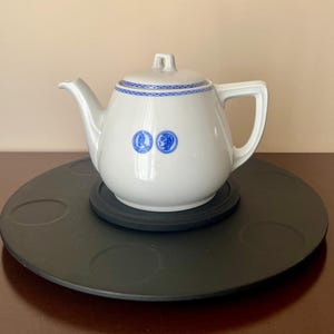May include: White ceramic teapot with a blue patterned border and two blue circular designs. The teapot has a curved spout and a handle. It sits on a black, round surface.