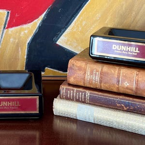 Op de afbeelding: Twee zwarte asbakken met een rechthoekige vorm en een rood en goudkleurig label met de tekst "DUNHILL". De asbakken zijn geplaatst op een stapel antieke boeken met bruine leren covers. De achtergrond toont een abstract schilderij.