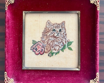 May include: A framed needlepoint featuring a fluffy cat with green eyes and a pink rose. The artwork is set in a gold-toned metal frame, mounted on a deep red velvet backing with decorative corner accents. A charming piece of vintage decor.