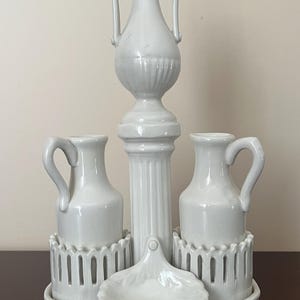 Może przedstawiać: Biały ceramiczny zestaw do przypraw z trzema pojemnikami i małą miseczką. Pojemnik środkowy to wysoki wazon z dwoma uchwytami. Dwa boczne pojemniki mają pojedyncze uchwyty. Zestaw jest ozdobiony wypukłym wzorem.
