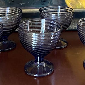 May include: A set of six vintage blue glass dessert bowls. Each bowl has a spiral design and a stemmed base. The glasses are arranged on a dark brown surface, with a painting in the background.