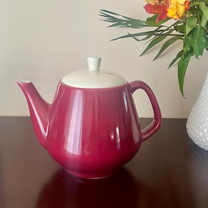 Puede incluir: Una tetera de cerámica burdeos brillante con tapa y asa color crema. La tetera tiene un cuerpo redondeado y un pico curvo. Un jarrón blanco con flores está en el fondo.