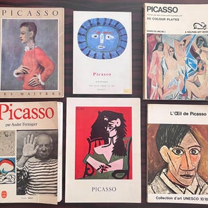 Może przedstawiać: Kolekcja starych książek o Pablo Picasso, z różnymi okładkami prezentującymi portrety i dzieła sztuki. Tytuły to m.in. "Picasso" i "Les Maitres". Książki ułożone są na ciemnej drewnianej powierzchni.
